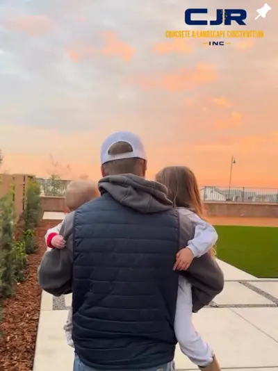 Colton Ray carrying two children across a new concrete walkway and artificial turf at sunset