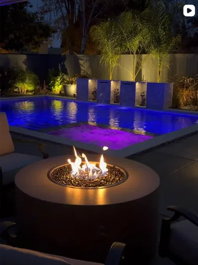 Round concrete fire pit lit at night beside a pool glowing blue and purple with three water features
