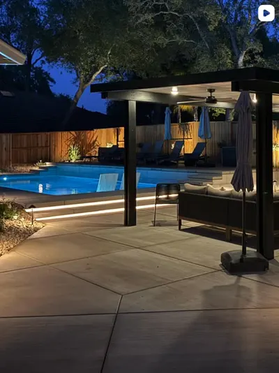 Covered patio with ceiling fan and outdoor seating beside a pool with step lighting at dusk